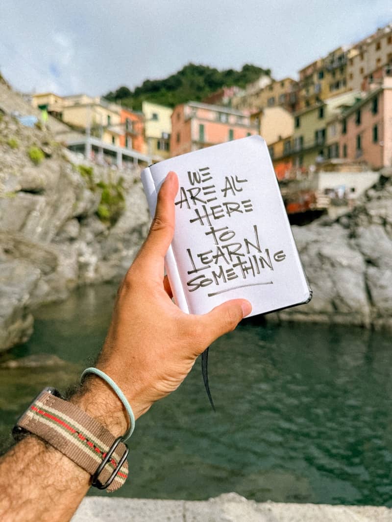 A hand holding a notebook reading "WE ARE ALL HERE to LEARN SOMETHING" by cliffs