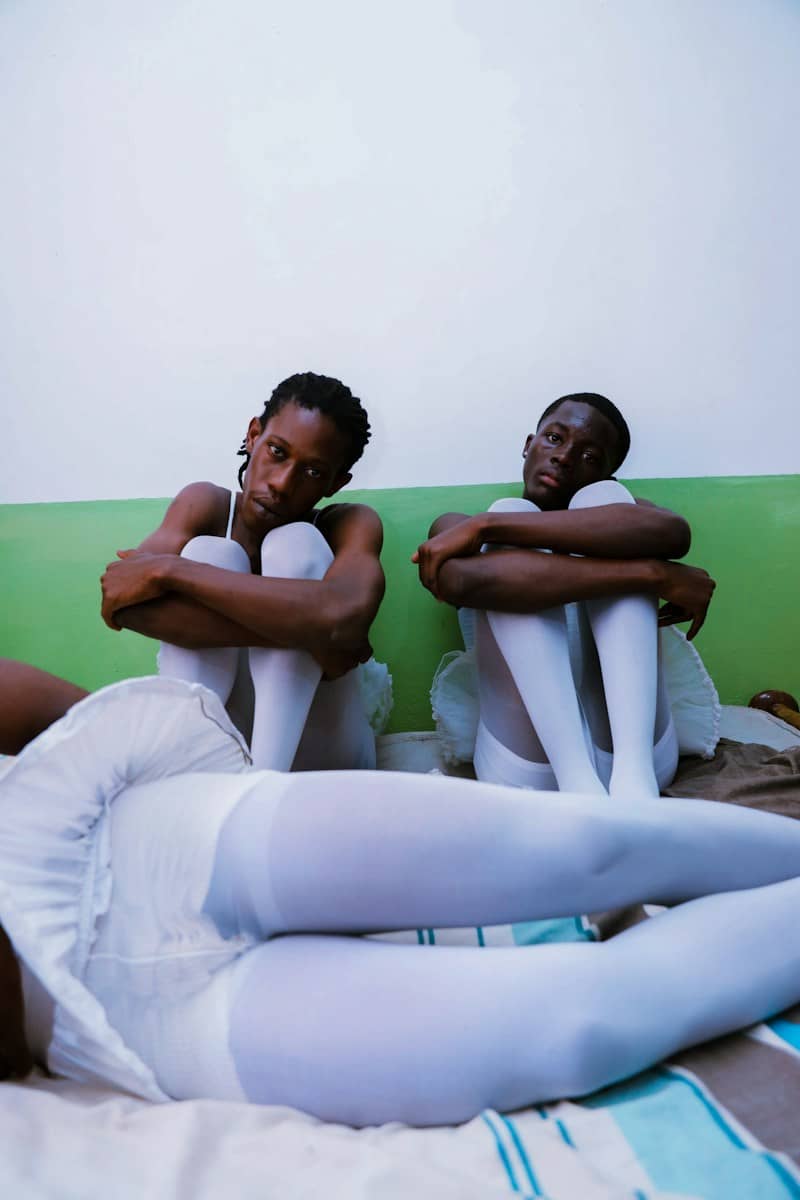 Three people in white ballet tutus and tights on a bed against a green wall