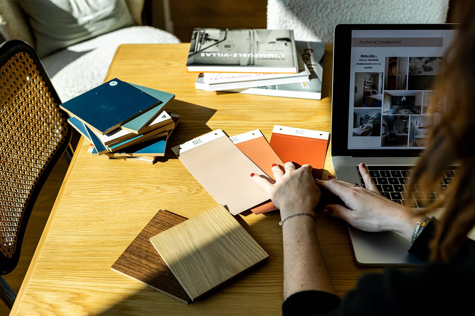 Main sélectionnant des échantillons de matériaux sur un bureau, à côté d’un ordinateur affichant une planche d’ambiance, illustrant la prise de décision et la responsabilité dans un projet professionnel.
