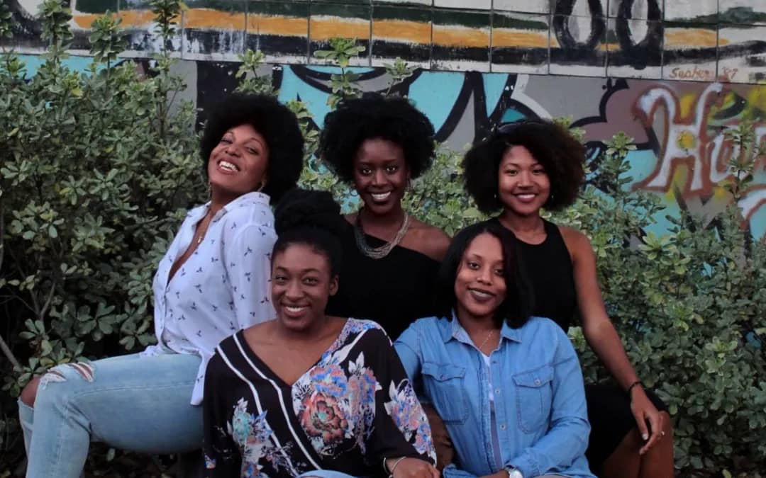 Five Black women sitting together in front of a mural, smiling and radiating joy, sisterhood, and confidence in a lush outdoor setting.