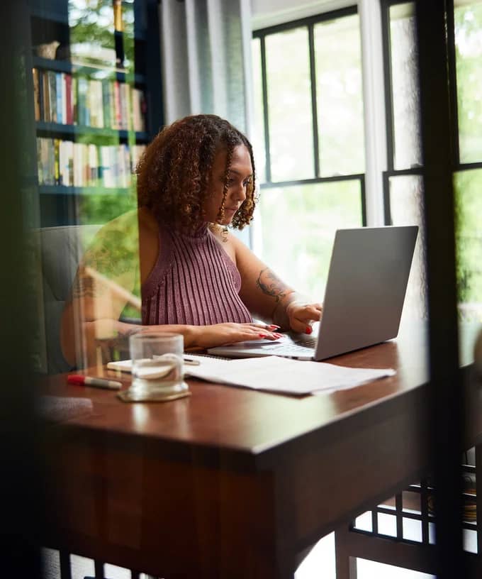 Woman with curly hair types on laptop at wooden desk near large window.