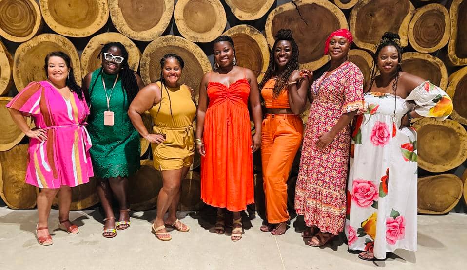 Seven women standing together in colorful dresses, smiling in front of a wood log wall backdrop.