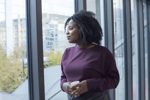 A thoughtful woman stands by a window, holding a cup and gazing outside with a calm, reflective expression.