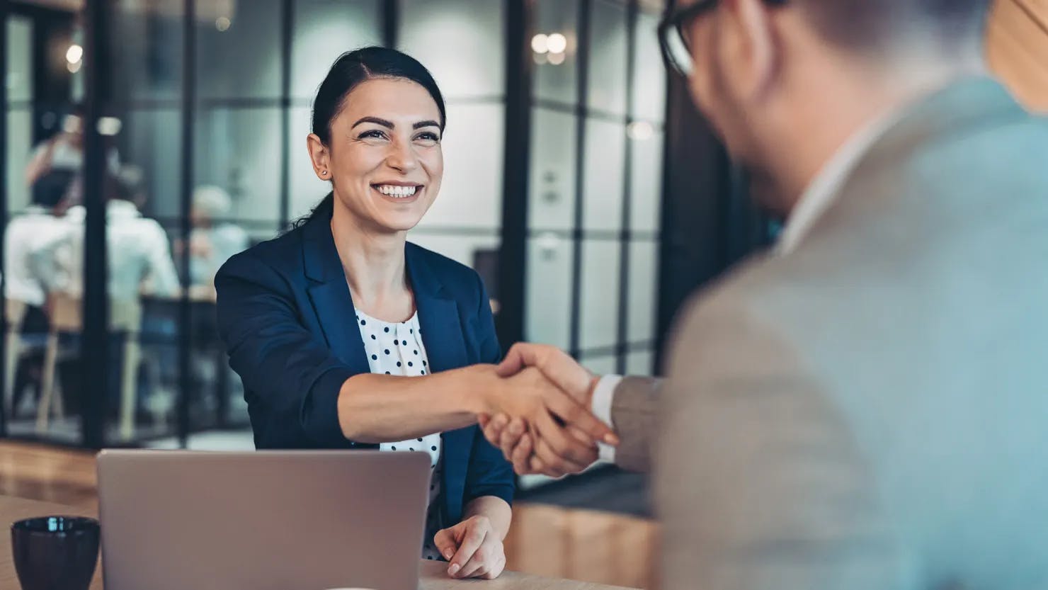 A man and a woman in business attire, smiling and shaking hands across a table.