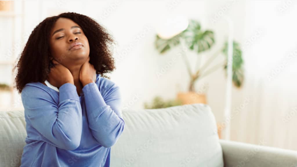 A tired woman sits on the couch, holding her neck in discomfort, showing signs of stress and fatigue.