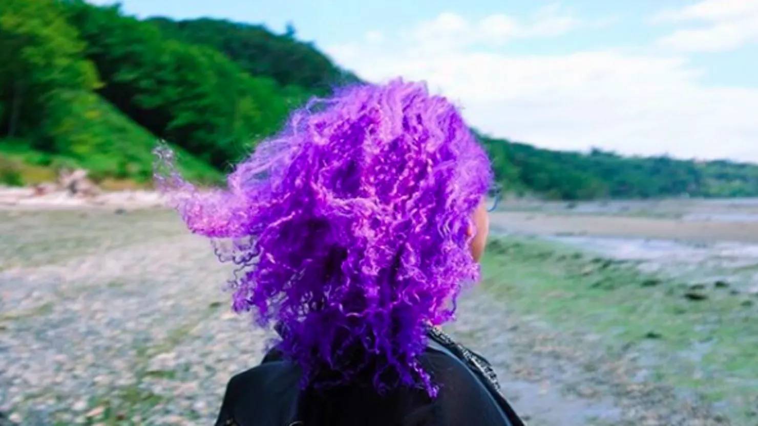 A Black woman with vibrant purple curly hair stands by a beach.