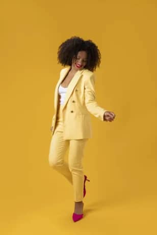 A joyful Black woman in a bright yellow suit and red heels smiles and dances confidently against a solid yellow background.