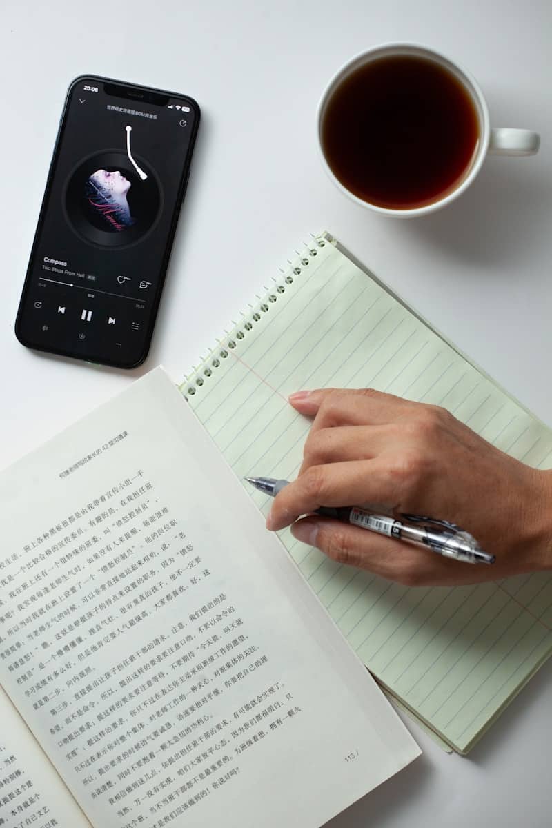 A person writing on a notebook next to a cell phone