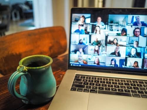 a laptop computer screen filled with zoom attendees beside a mug next to a wooden chair and a window overlooking a suburban scene
