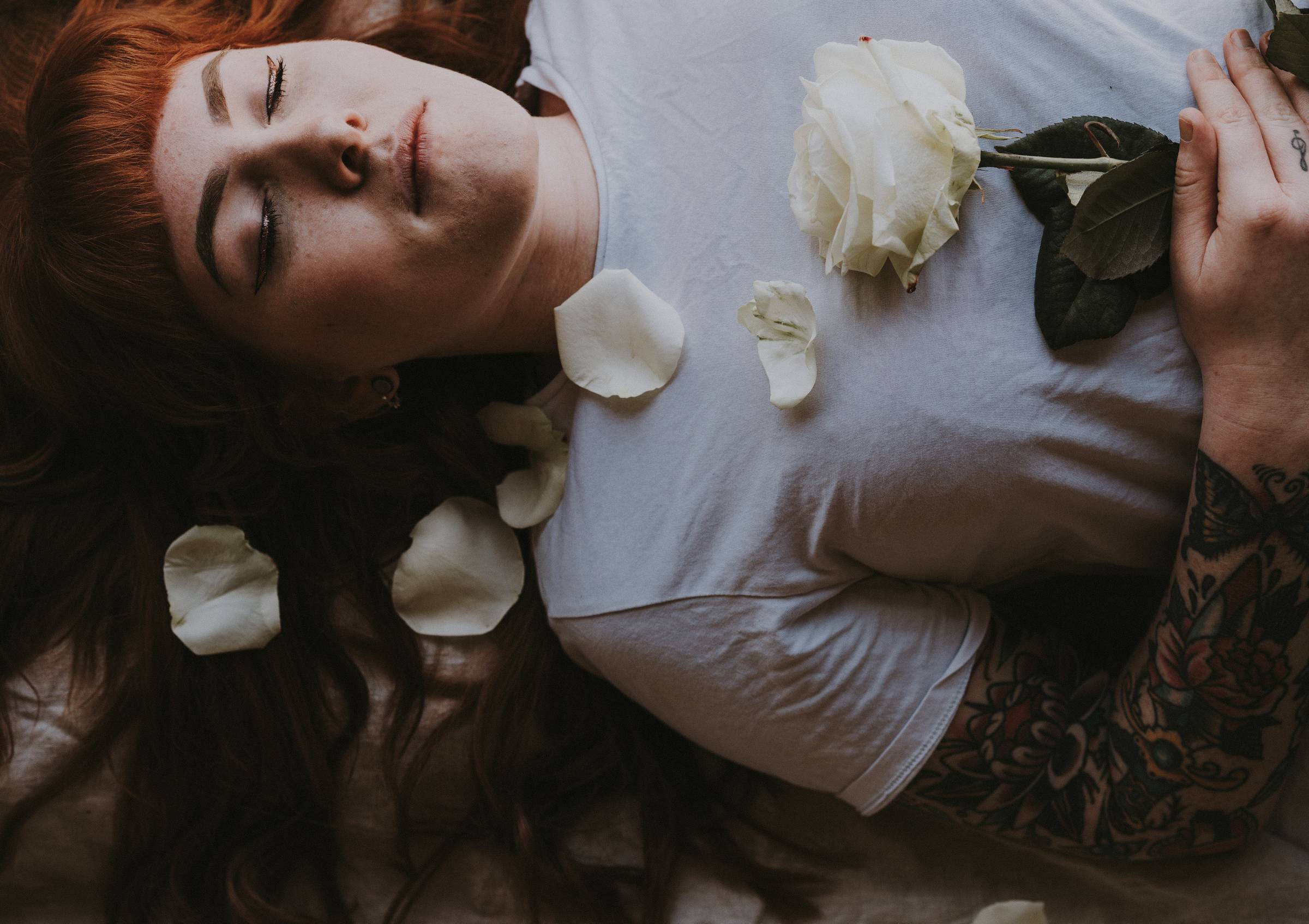 A woman laying on her back wearing a white tshirt and holding a white rose, with petals falling around. Her eyes are closed.