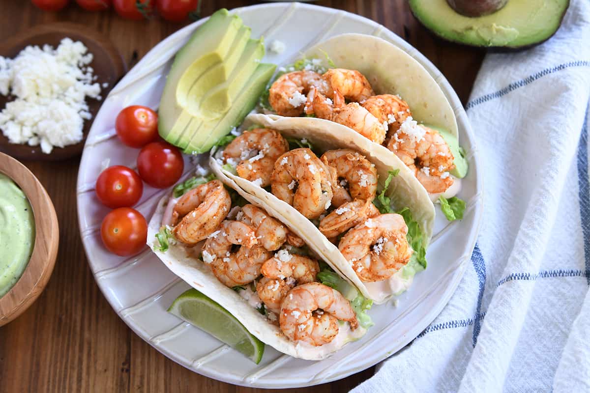 A plate of three shrimp tacos garnished with crumbled cheese and lettuce. The tacos are accompanied by slices of avocado, cherry tomatoes, and a lime wedge on the side. A small bowl of green sauce and crumbled cheese are visible in the background, with half an avocado and a checkered cloth nearby.