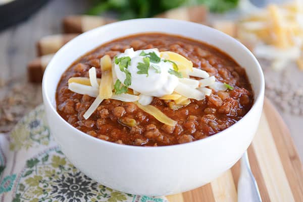 A bowl of chili topped with shredded cheese, a dollop of sour cream, and a sprig of cilantro. The chili appears hearty, with a rich, red sauce, and is set on a wooden board with a patterned cloth beneath it. There is a spoon placed beside the bowl.