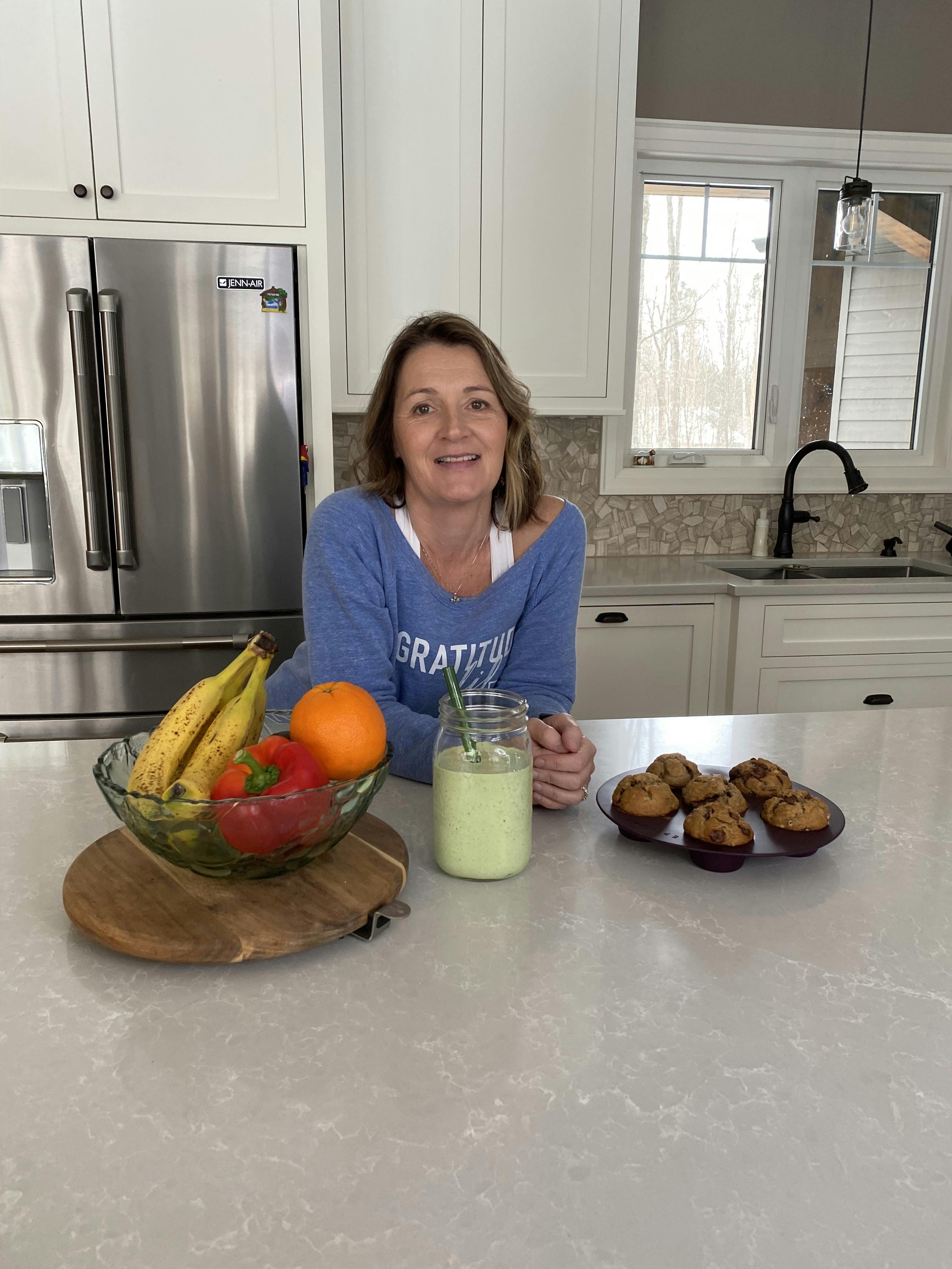 Anita Perrigo leaning onto her kitchen island with a green smoothie, a bowl with fruit and vegetables and fresh muffins