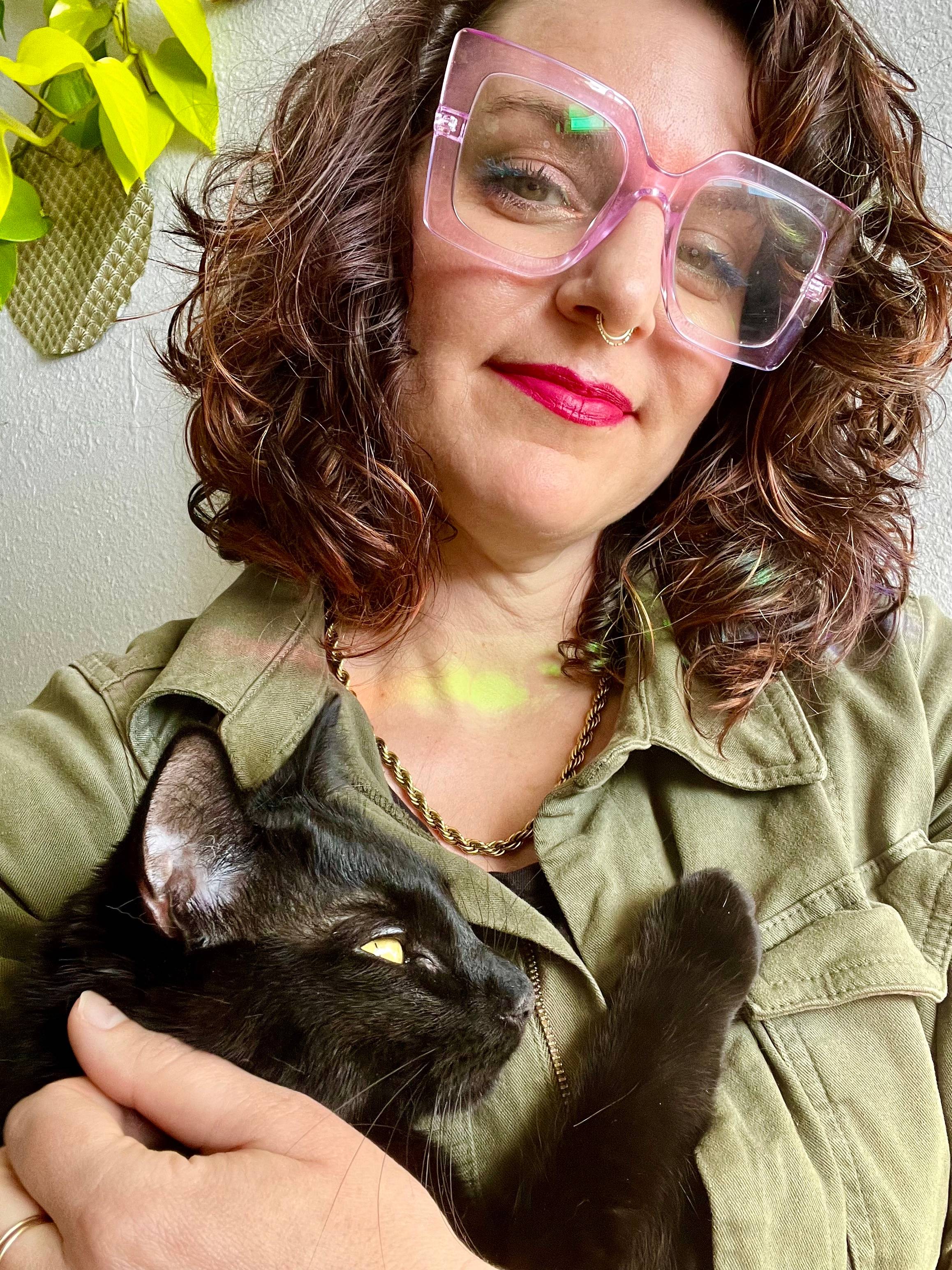 a brunette wearing big square purple glasses, with curly hair to their shoulders smiles in pink lipstick, tilting their head to the side smiling at the camera while holding a black cat with his paw on their chest