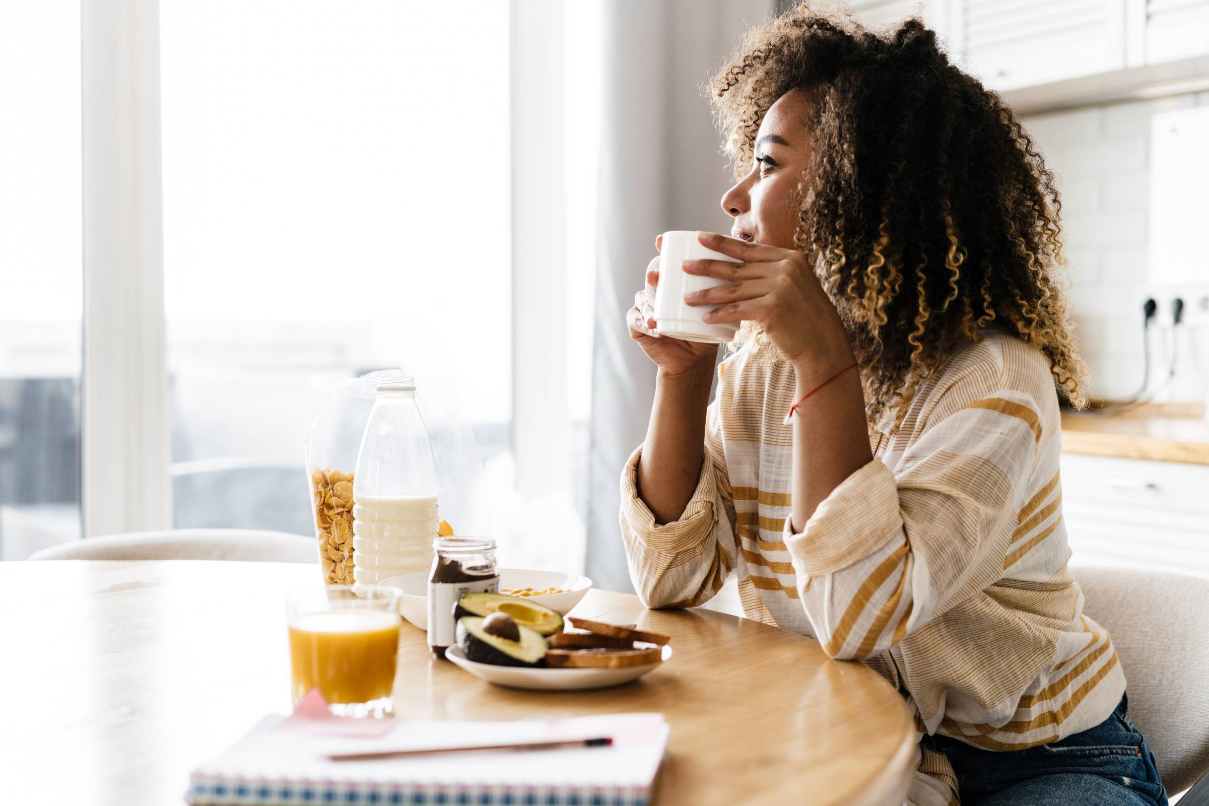 The side view of a smiling woman holding a cup in her hands and looking out the window while sitting in the kitchen