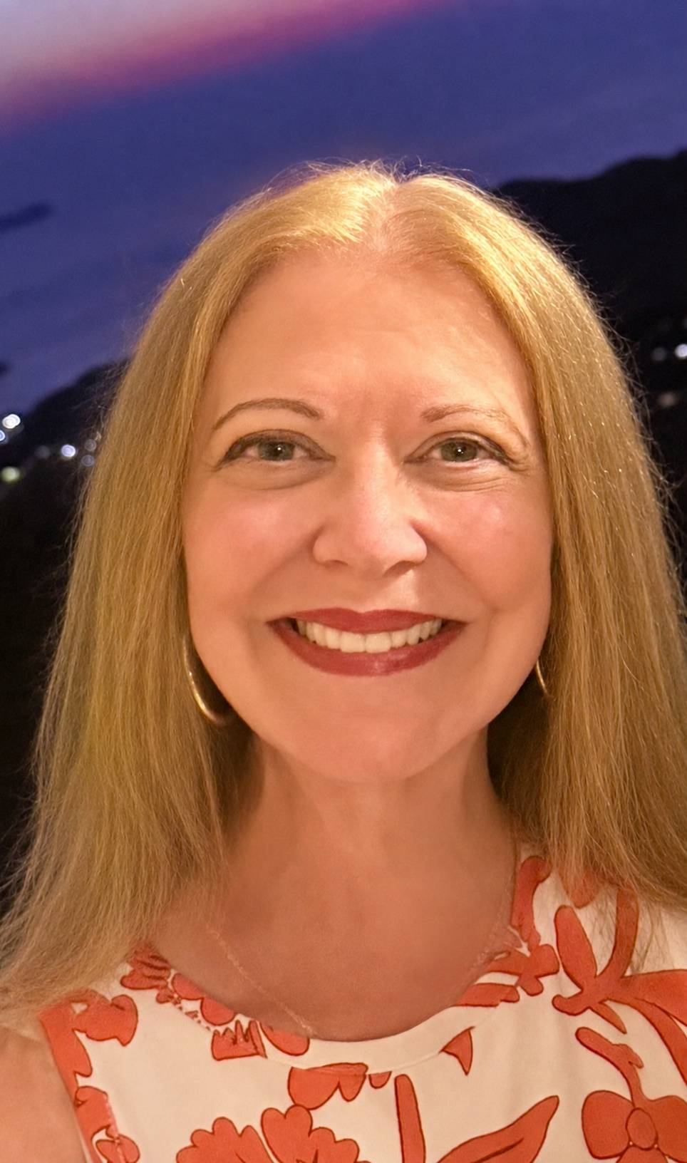 Cari smiles at the camera, wearing an orange and white floral top and gold hoop earrings.