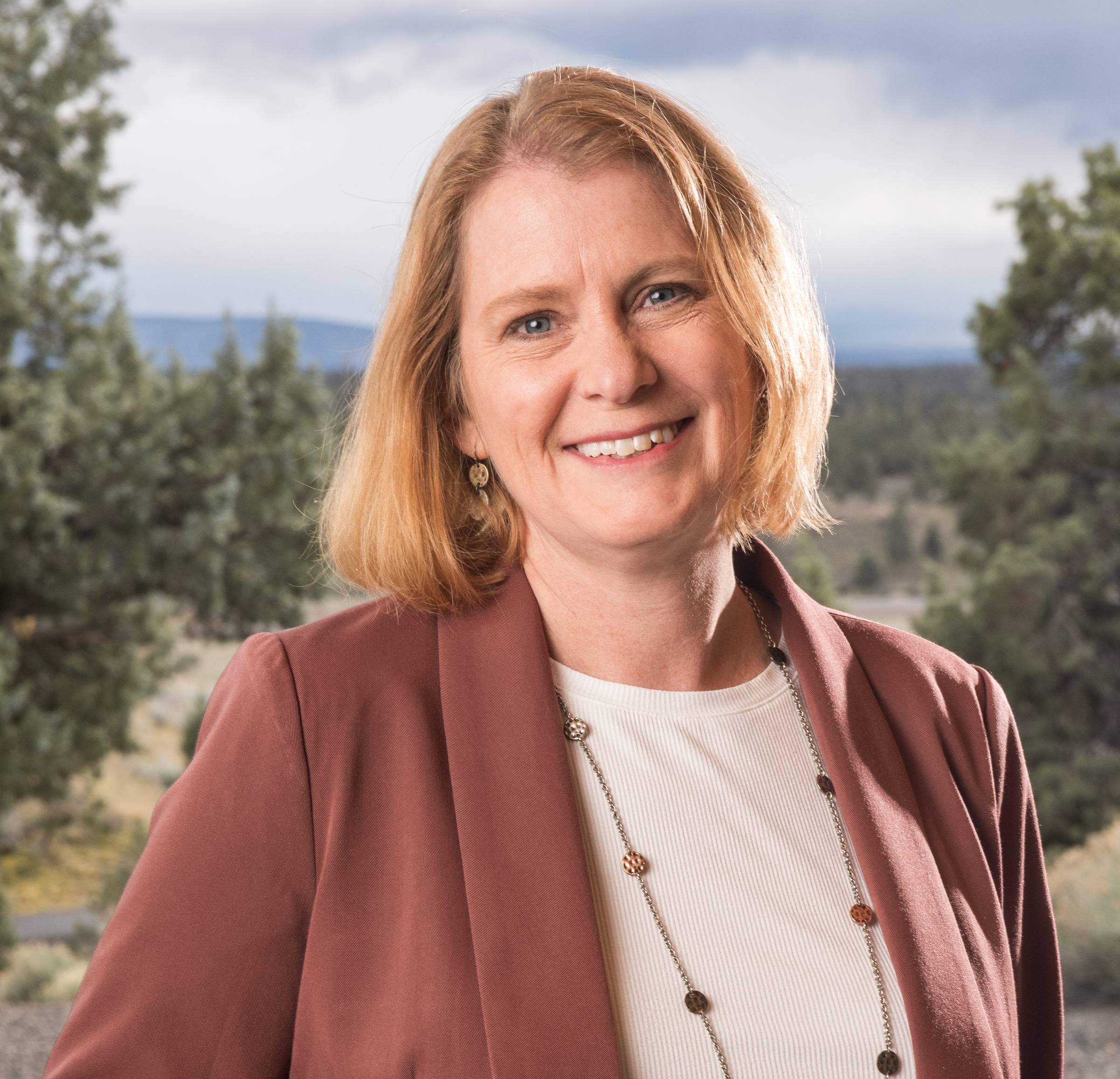 Ashley Feit's headshot. She wears a dark mauve blazer over a cream blouse with a beaded necklace and beams in front of a forest landscape in the horizon.