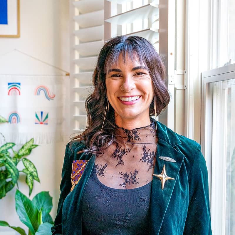 Jen's headshot. She smiles at the camera, wearing a green velvet blazer with assorted broaches and a black lace blouse.