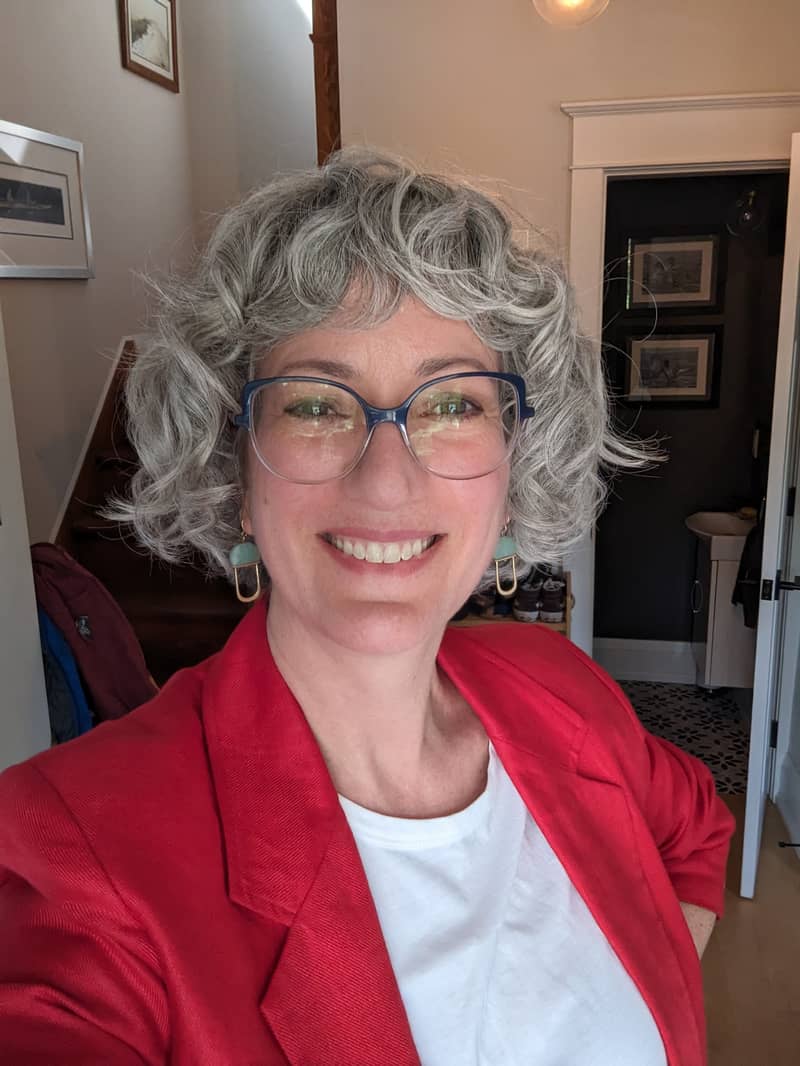 A photo of a smiling white woman with wavy silver hair, wearing glasses and a red blazer.
