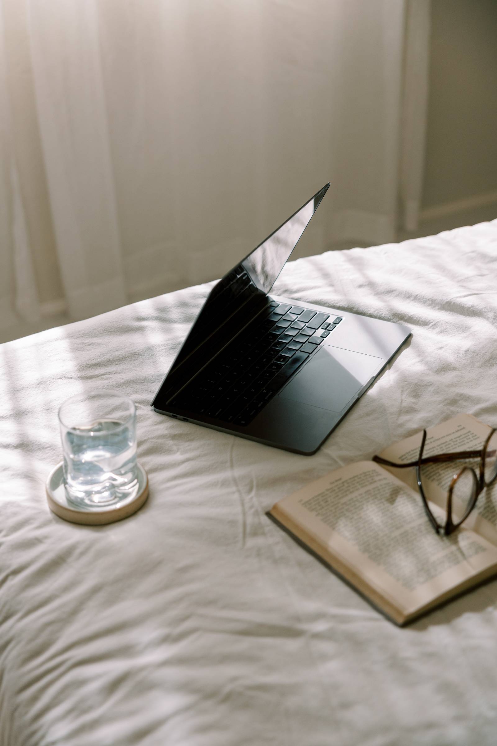 Image of a laptop, book, glasses, and for weird reason a glass of water on a bed (highly impractical)
