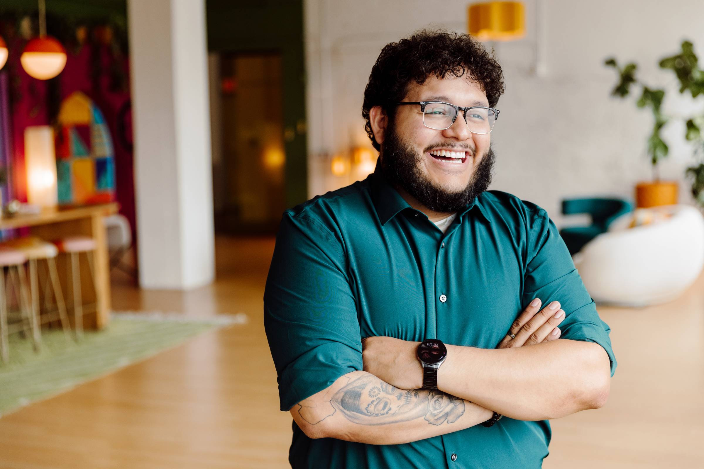 A person with short curly hair, a beard, and glasses stands smiling with arms crossed in a brightly lit, modern indoor space. They are wearing a dark green button-up shirt and a black wristwatch, with a visible tattoo on their forearm. The background includes colorful artwork, plants, and cozy furniture. 