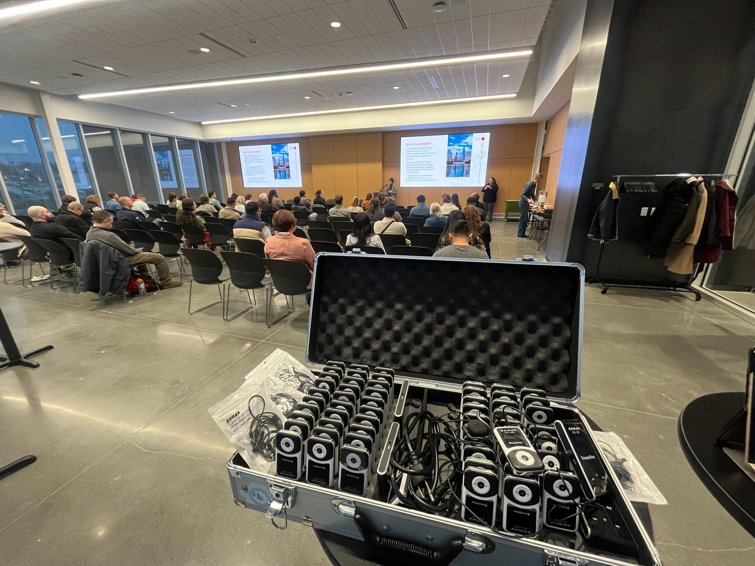 A box of interpretation devices sits in a room full of attendees. In front of them are sign language interpreters and screens. 