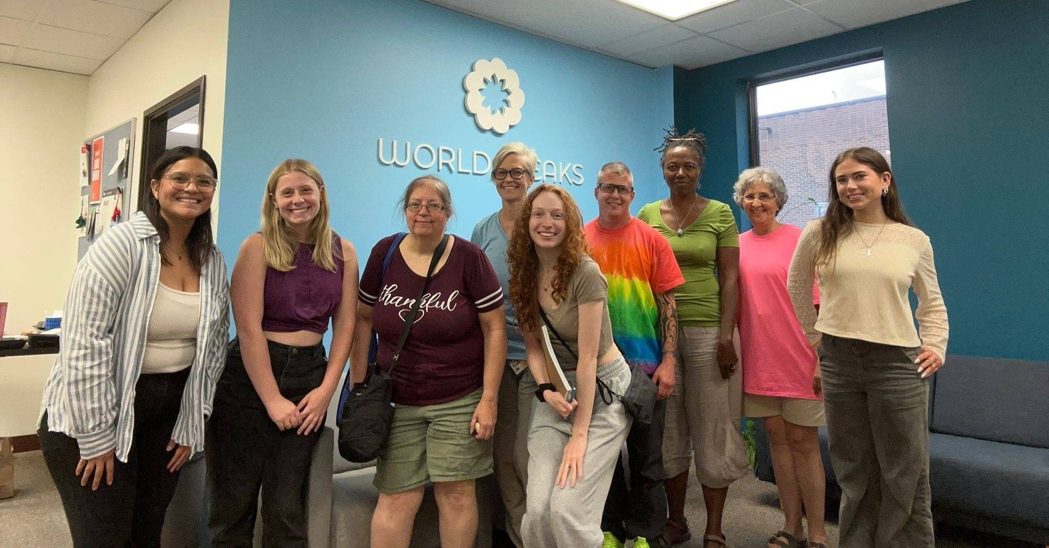 A group of ten people stands smiling inside the World Speaks office. They are casually dressed and standing in front of a blue wall with the World Speaks logo. The room is well-lit with natural and fluorescent light, and a window shows part of a brick building outside.