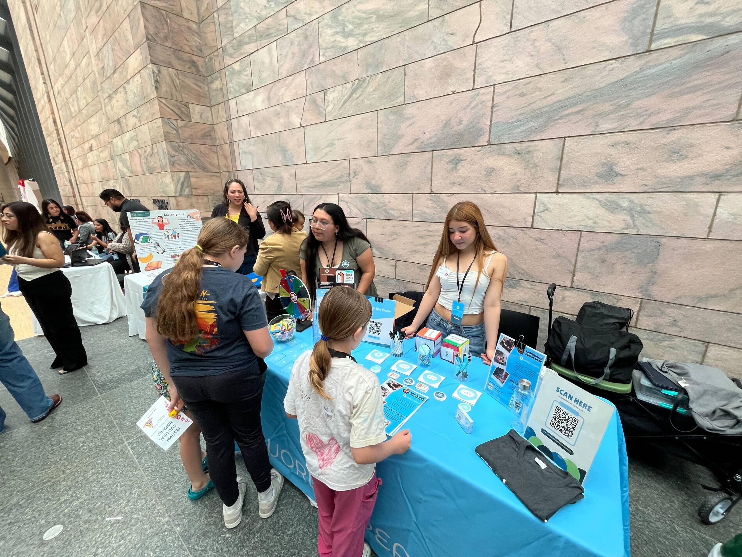 Two young women staff a bright blue exhibition table at an indoor event, engaging with visitors including two children. The table displays informational materials, QR codes, and small giveaway items. Other exhibitor tables are visible in the background.