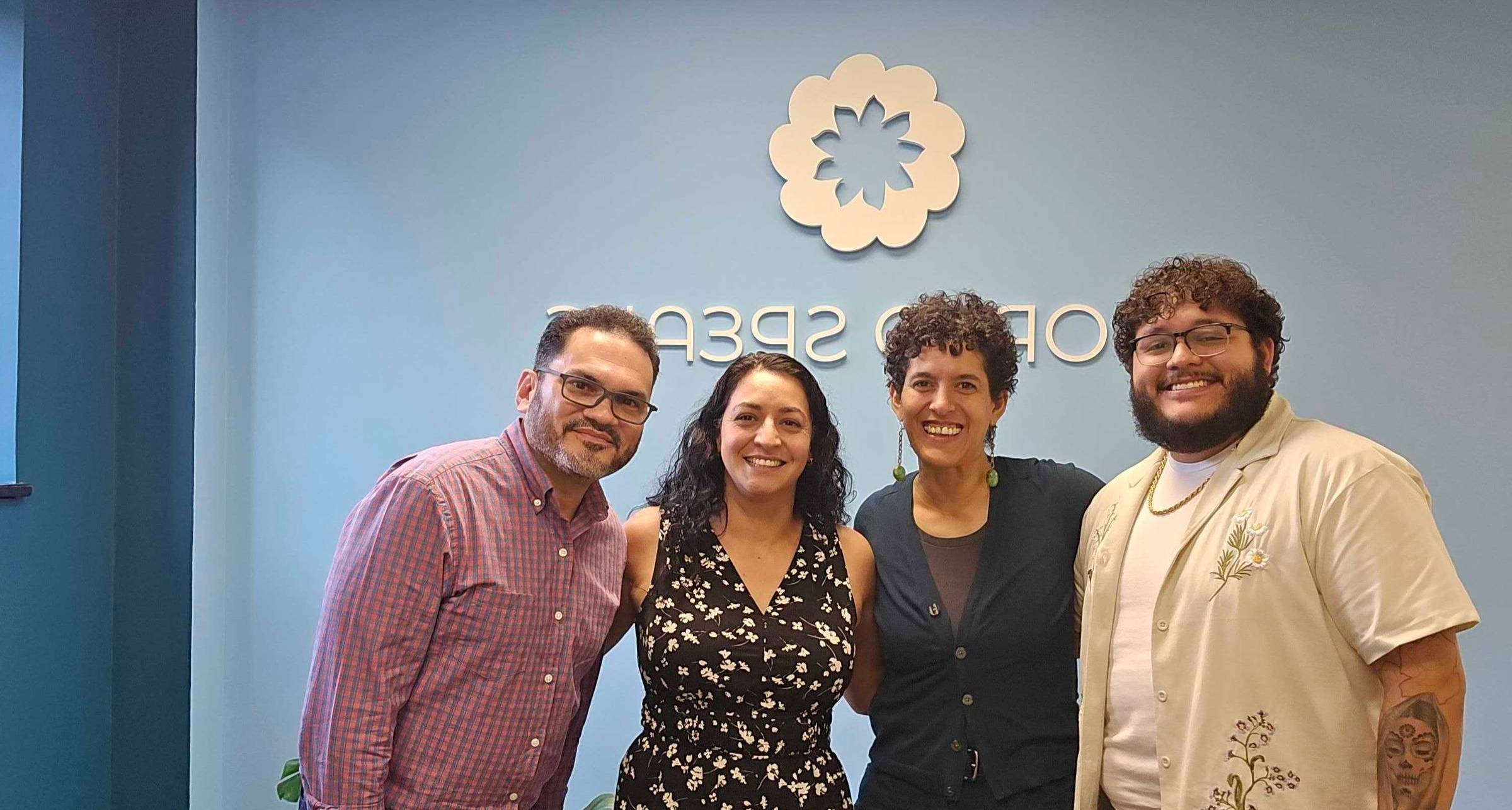 Four people stand closely together, smiling, in front of a light blue wall with the World Speaks logo. The group includes two men on the left and right and two women in the middle. They are standing in an office space with couches and plants visible in the background.