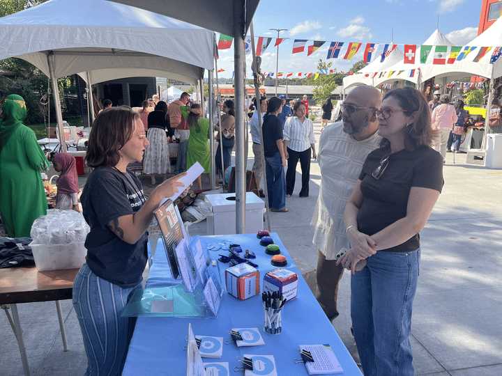 Three people stand at a display table at a festival. One reads some information to the other two. Colorful flags and crowds stand behind them. 