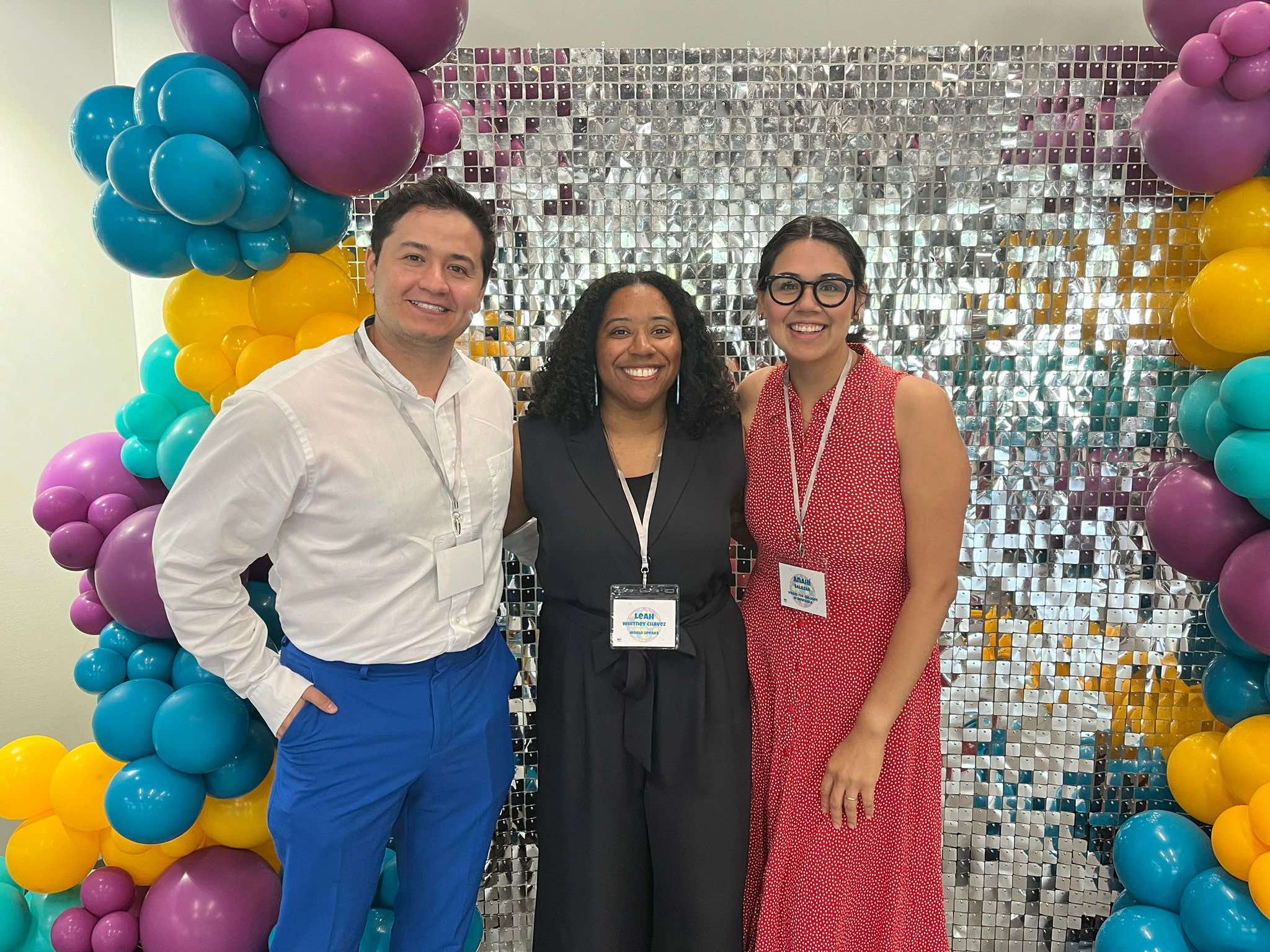 Three people stand in front of a festive backdrop. They are all smiling at the camera.