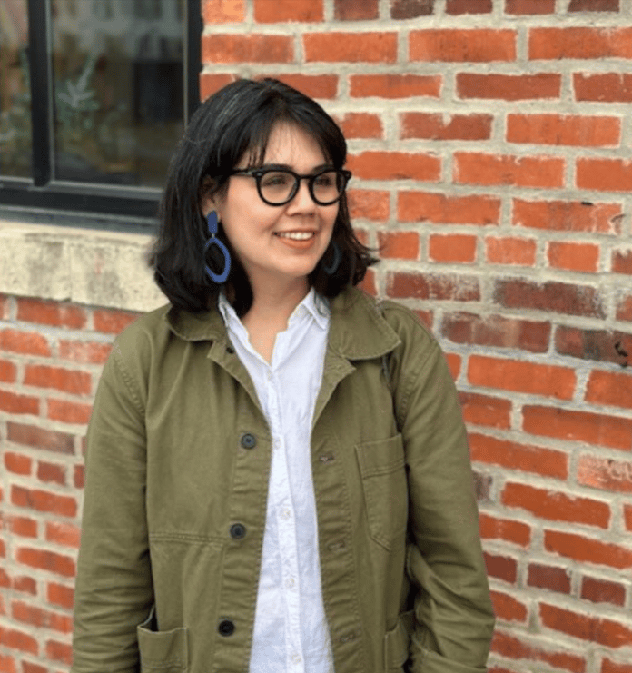  Image shows a woman with dark hair, glasses and bright blue earrings wearing a green jacket, standing in front of a brick wall. She is looking off to the side of the image, rather than at the camera.