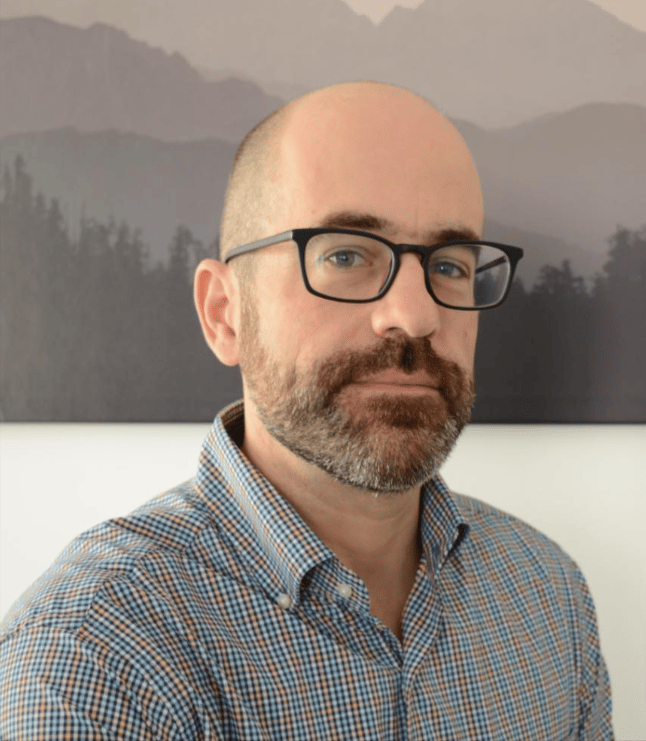 Image shows a man with a beard and glasses standing in front of a mural. He looks directly at the camera.