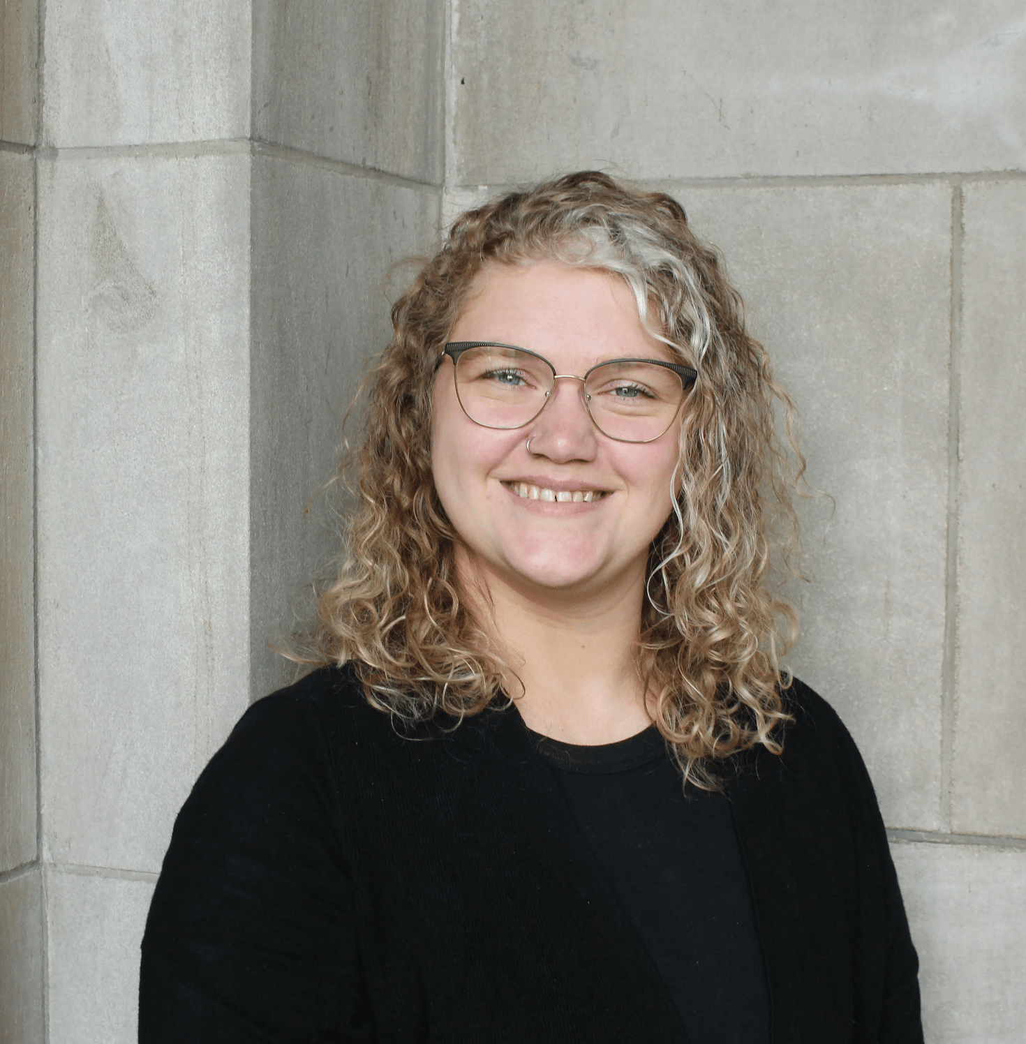 A blonde woman with curly hair, wearing glasses, wears a black t-shirt and smiles at the camera. 