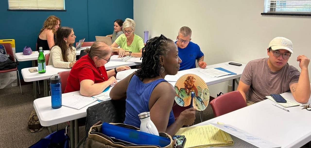 A group of people sit in a classroom. They are talking to teach other and working on worksheets.