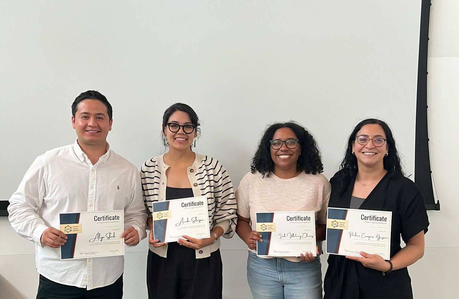 Four people stand in front of a white wall. They are all holding certificates and are smiling at the camera. 