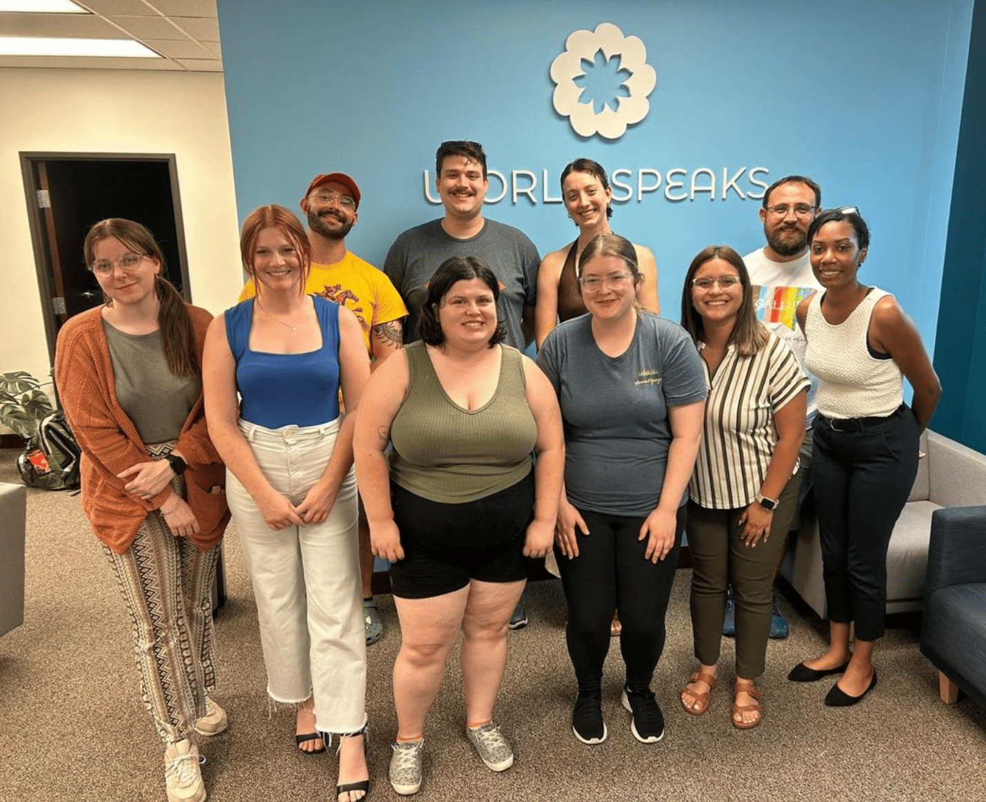 Image shows a group of people standing in front of a blue wall. The World Speaks logo is behind them.