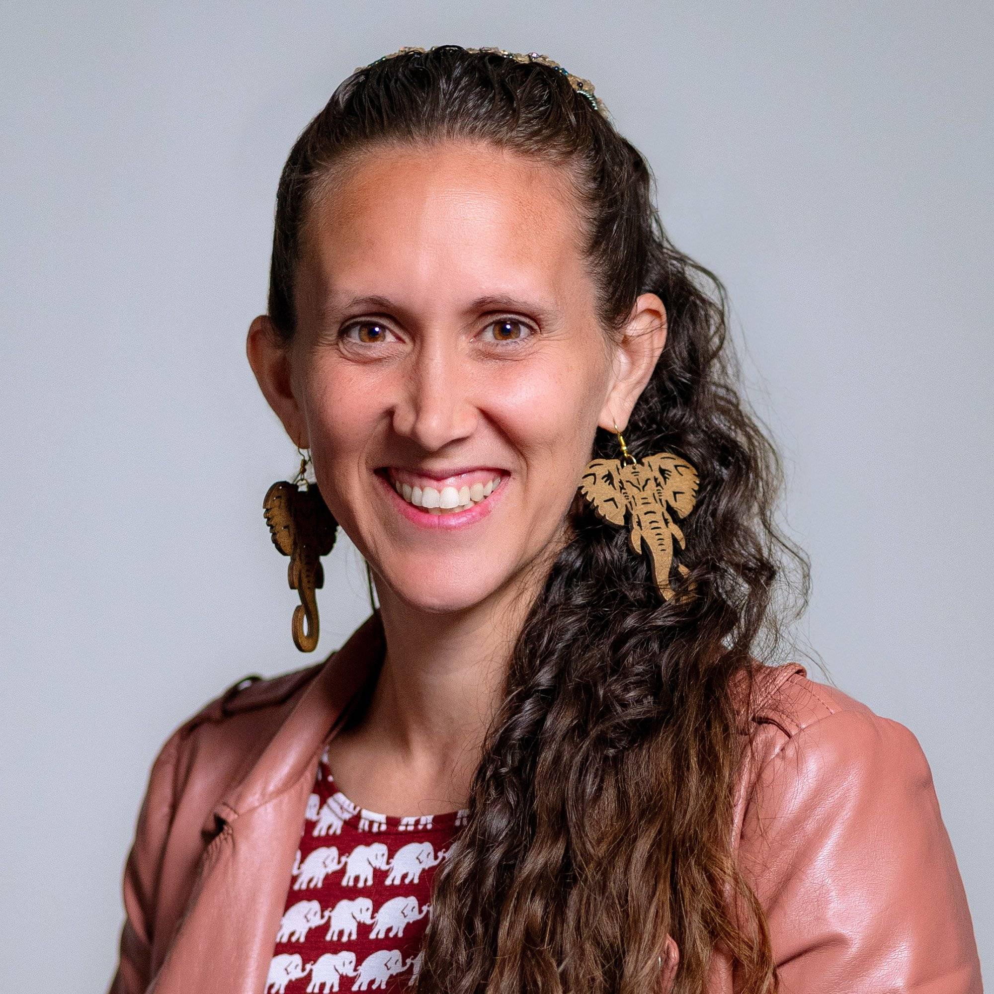 A smiling person with long brown hair and large earrings smiles at the camera in front of a grey background. 