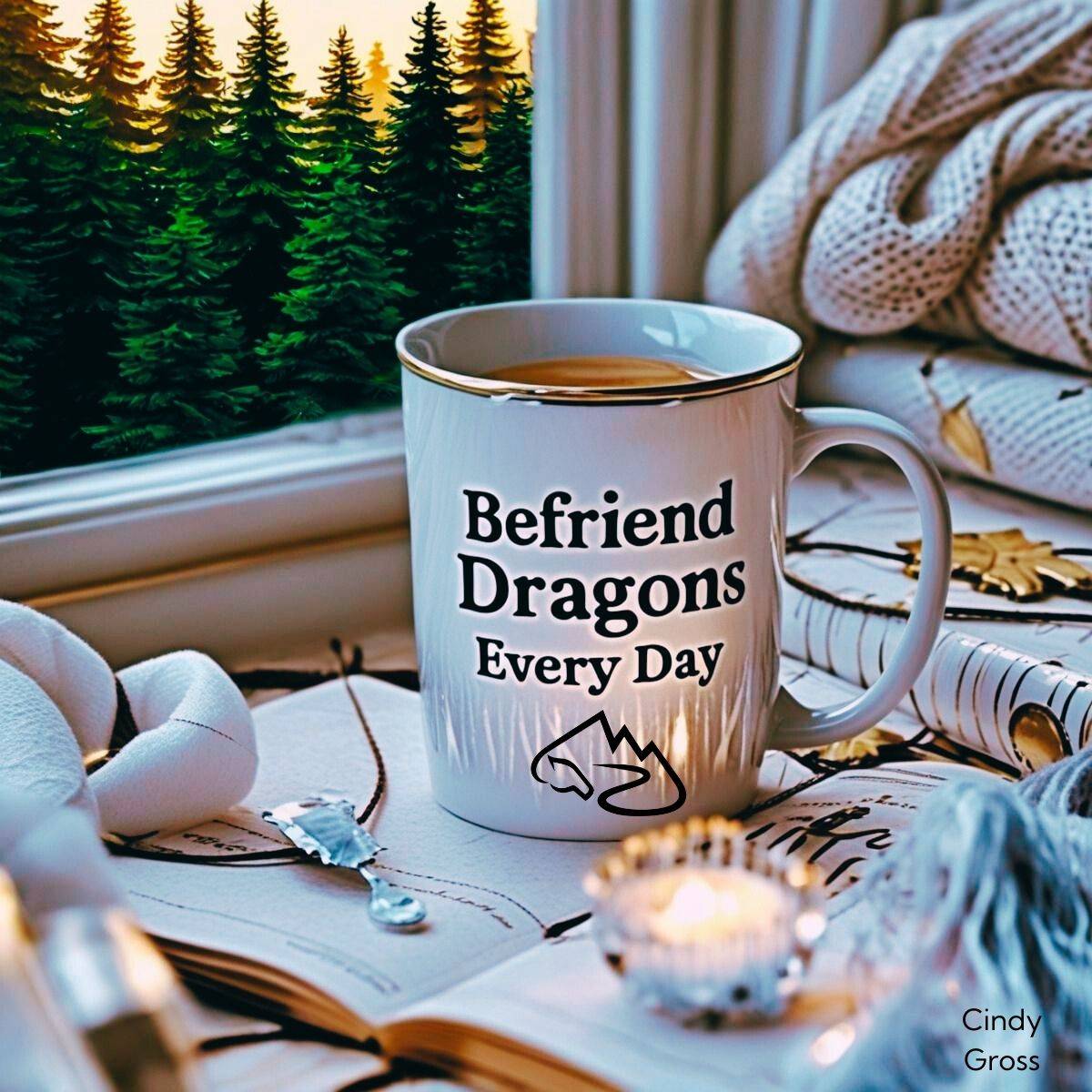 White and gold coffee mug with text "Befriend dragons every day" - sitting near a candle, a stack of journals, and a cozy white sweater in front of a window overlooking pine trees.