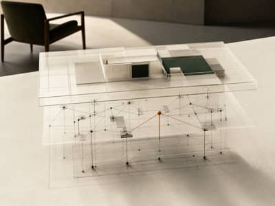 A translucent architectural model on an ivory table reveals hidden support lines beneath an intact surface, with one cognac load path and an empty chair in the background.