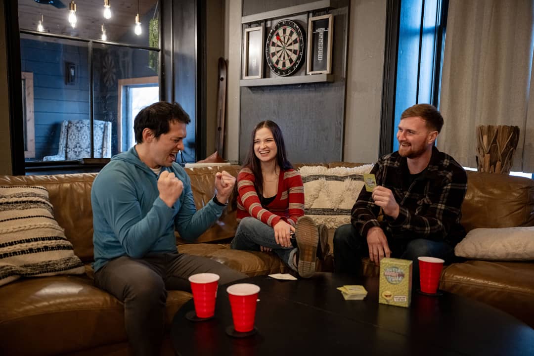 A group of three friends playing Not The Brightest Bulb in a cozy living room. The player in the blue hoodie is cheering triumphantly, while the player in the red-striped sweater laughs and the player in the black flannel grins while holding a card. The game box and prompt cards are visible on the table alongside red plastic cups, creating a lively and engaging game night atmosphere.