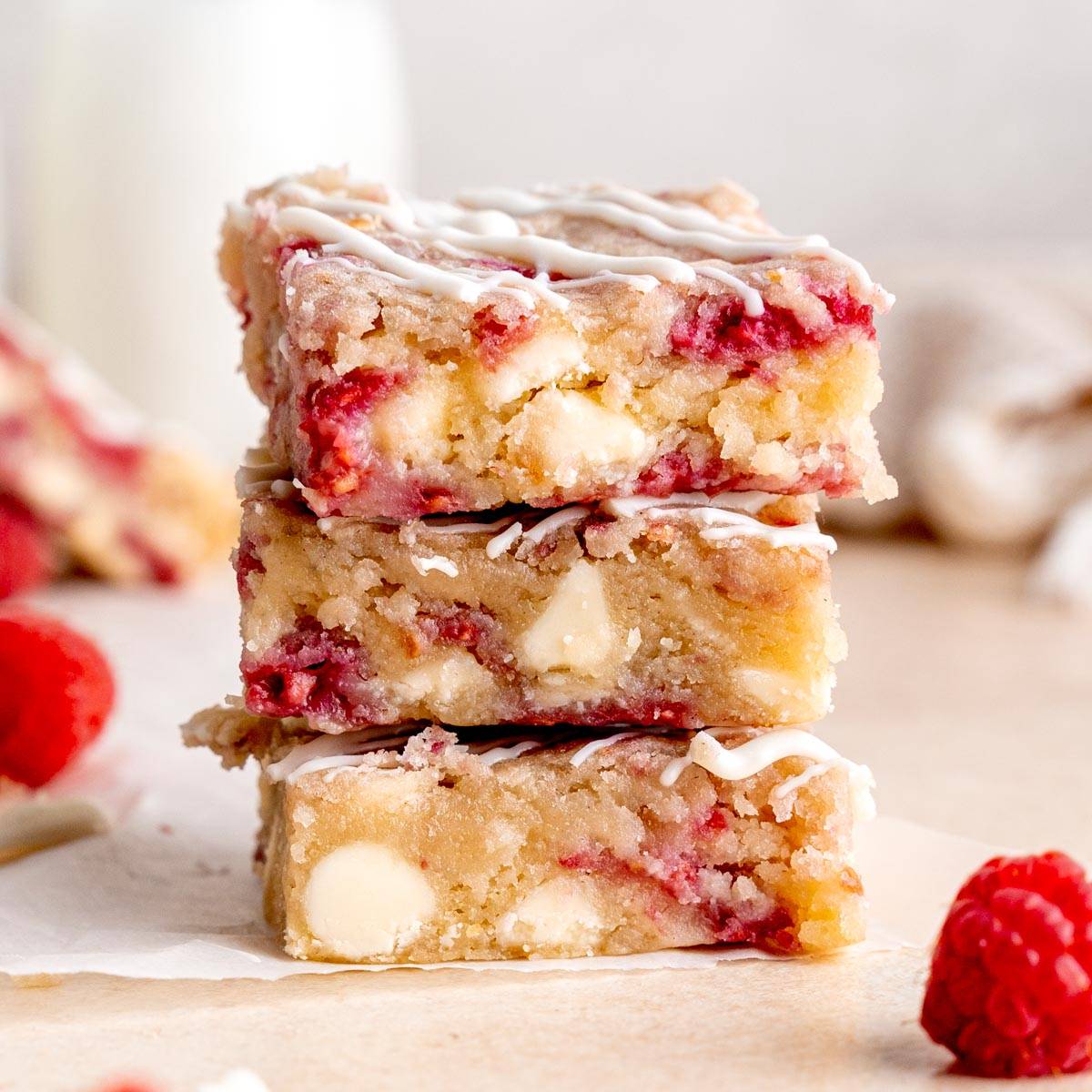 A stack of three raspberry and white chocolate blondies sits on a piece of parchment paper. The blondies are golden brown with visible red raspberry swirls and white chocolate chunks. A drizzle of white icing is on top of each bar. Fresh raspberries are s