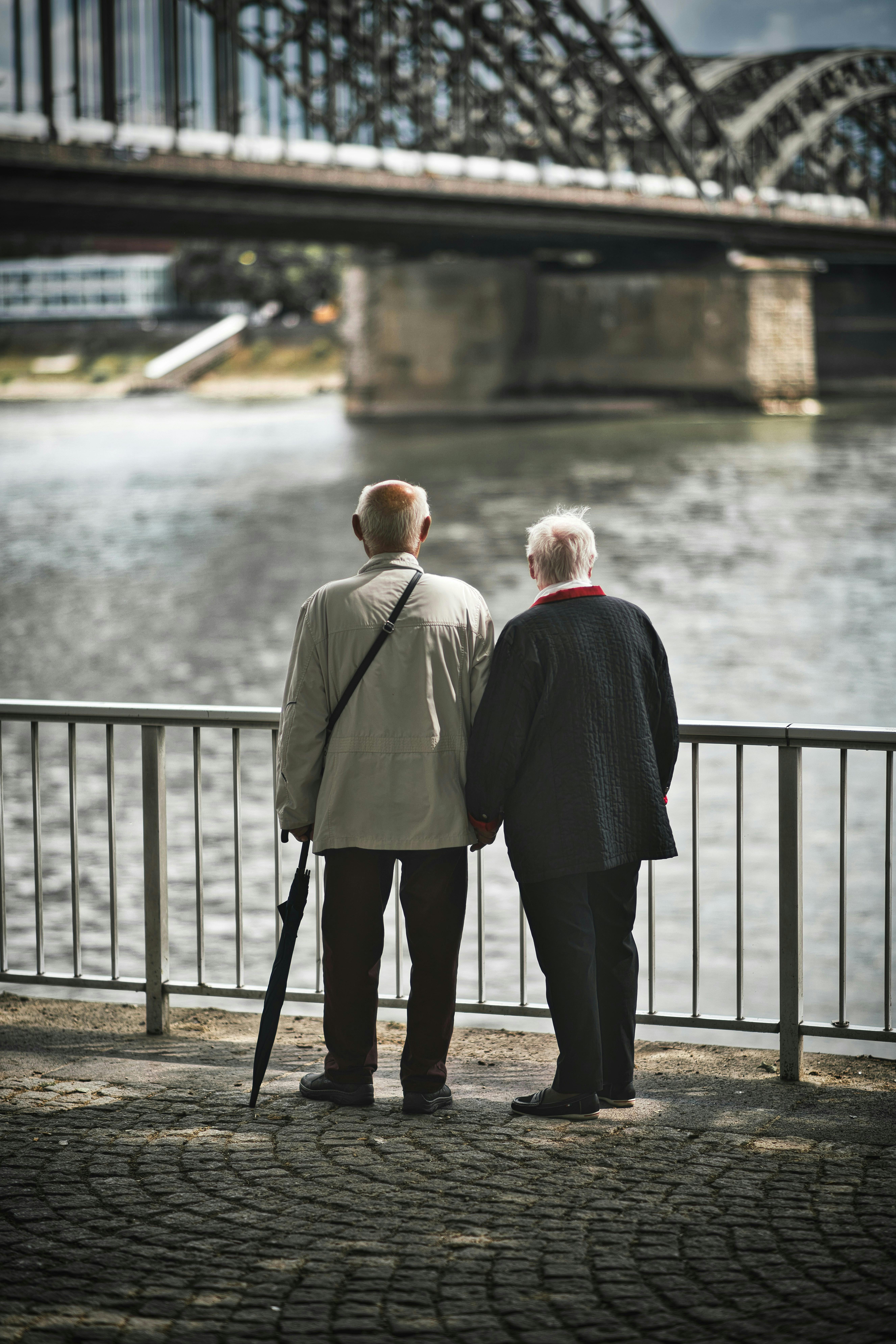 An older couple, viewed from the back, are holding hands while looking at a bridge over a river.