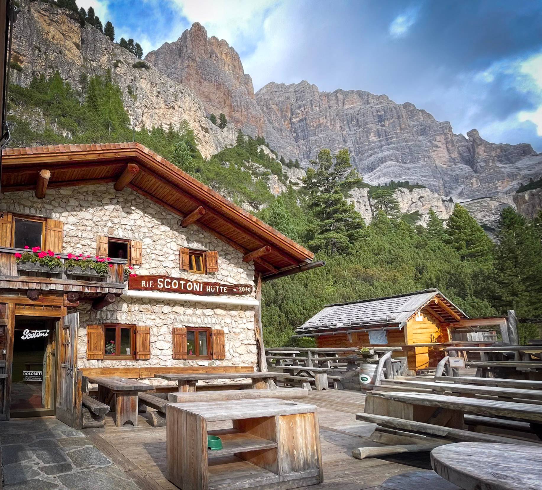 A building with a stone facade and wooden roof and tables out front set against a dramatic mountain background