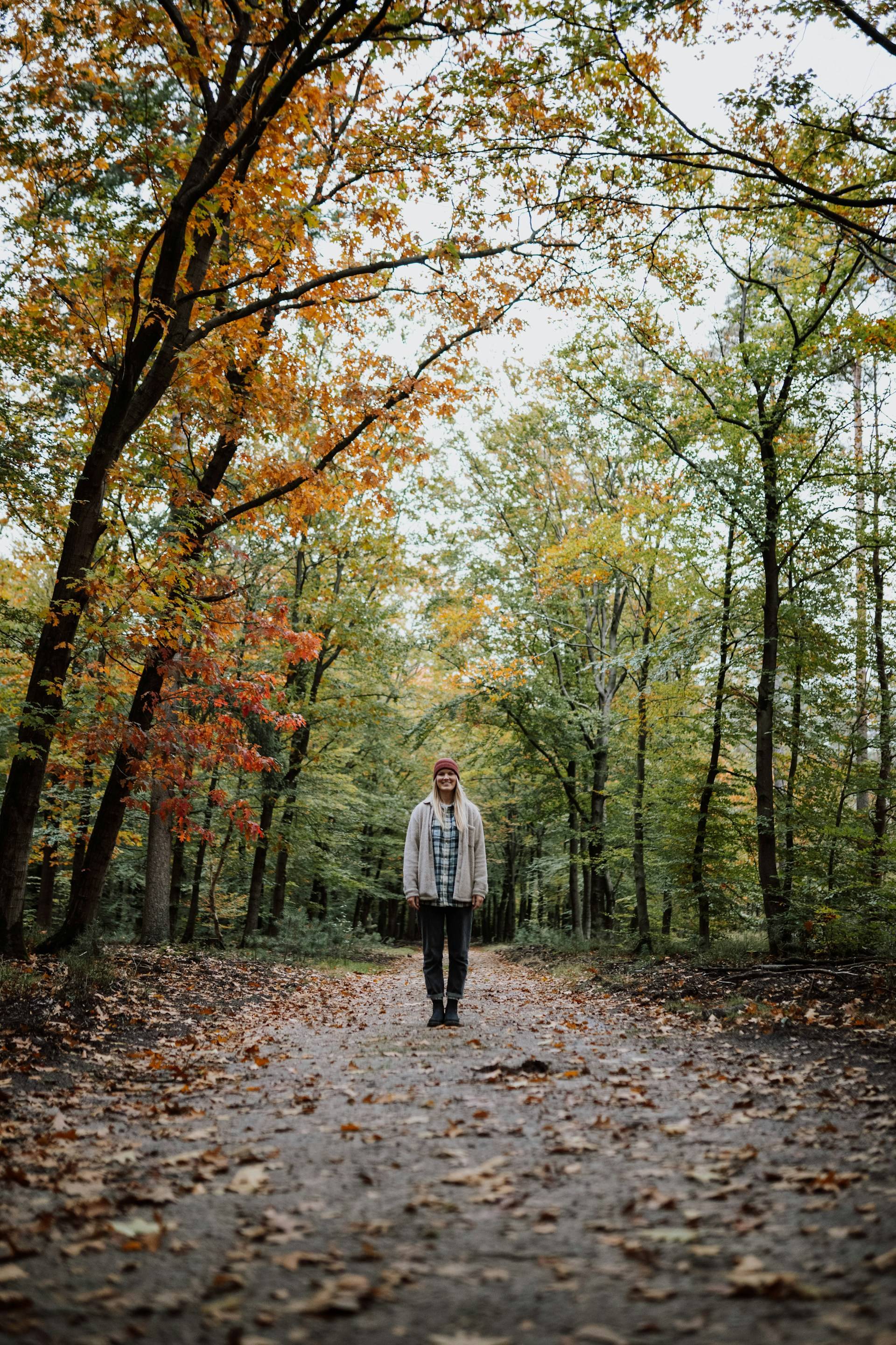 A midlife woman standing on a fall walking trail amidst the trees.