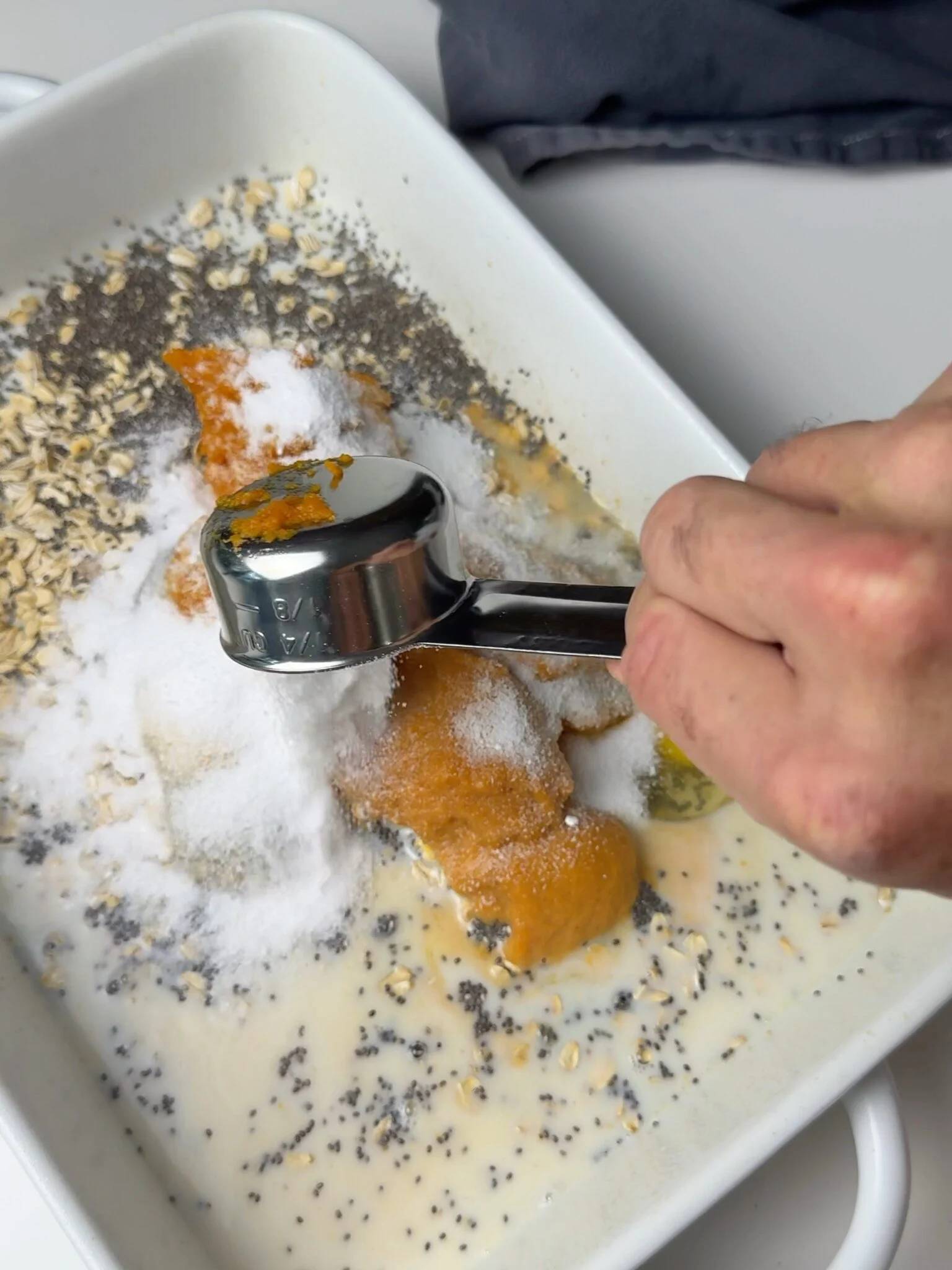 A measuring cup adding ingredients to pumpkin-spice baked oats in a white baking pan
