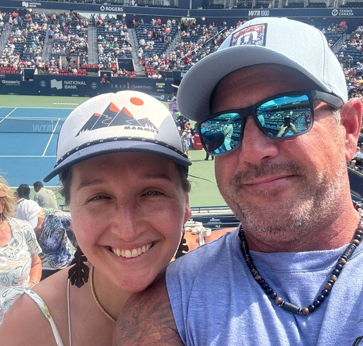 Tarzan and boyfriend taking a selfie from the stands at a professional tennis match, with the tennis court and crowd visible behind them.