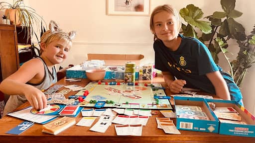 Tarzan's kids smiling while playing a colorful board game (Monopoly) together at a dining table surrounded by cards, game pieces, and supplies.