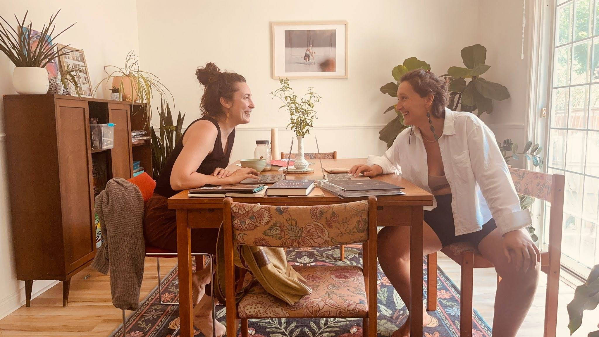 Tarzan Kay and Kendra Hoffman collaborating at a dining table with laptops, smiling as they work together on values-driven business strategy and creative planning in a bright home office.