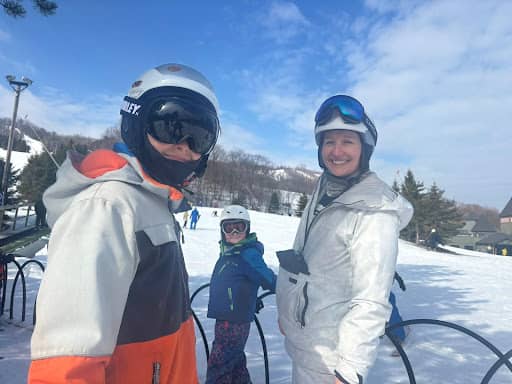 Tarzan and her son in ski helmets and goggles smile at the camera on a snowy ski slope; one wears a gray-and-orange jacket and the other a white snowsuit. Her youngest son in a blue jacket and helmet stands behind them near a metal railing, with snow-cove